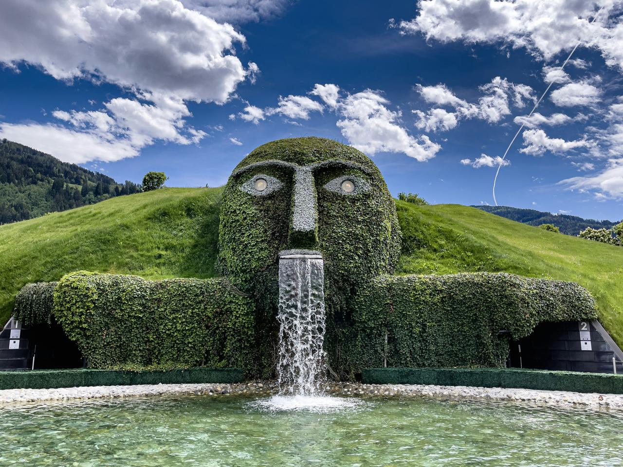 Große, grüne Skulptur eines Gesichts mit Wasserfall, die in eine grüne Wiese eingebettet ist