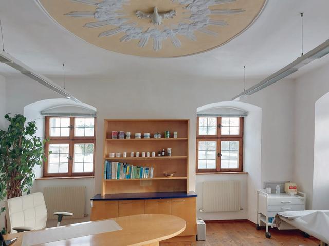 Ein helles, modernes und gut ausgestattetes Zimmer, das möglicherweise als Pflege- oder Therapiebereich dient, mit einem auffälligen, sonnenstrahlartigen Deckenkunstwerk.